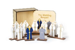A wooden nativity scene set titled "The Wedding Banquet" featuring painted wooden figures in white robes with a blue-robed central figure, arranged in front of a wooden box.